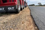 A B-double truck parked on the dirt shoulder of a highway with the bitumen crumbling on the edges.