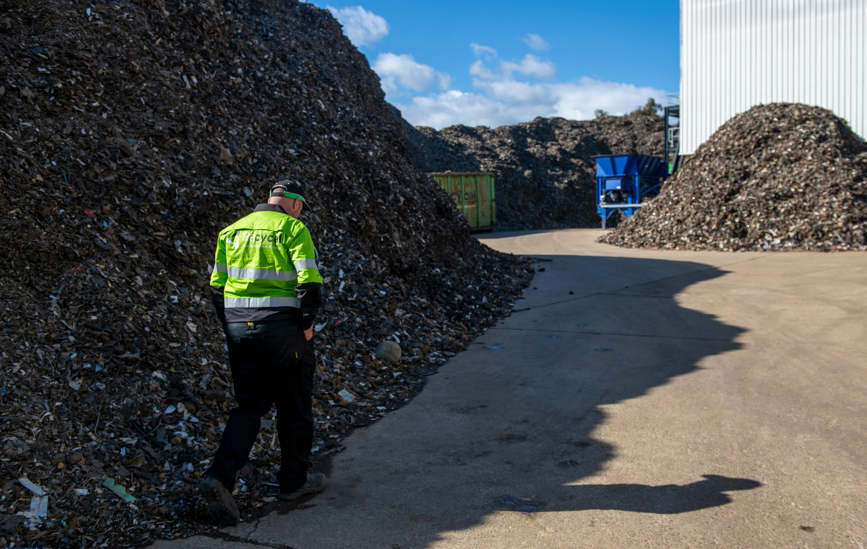 A man in a fluro yellow vest walks passed large piles of shredded scrap metal, a green shipment container and blue machinery.