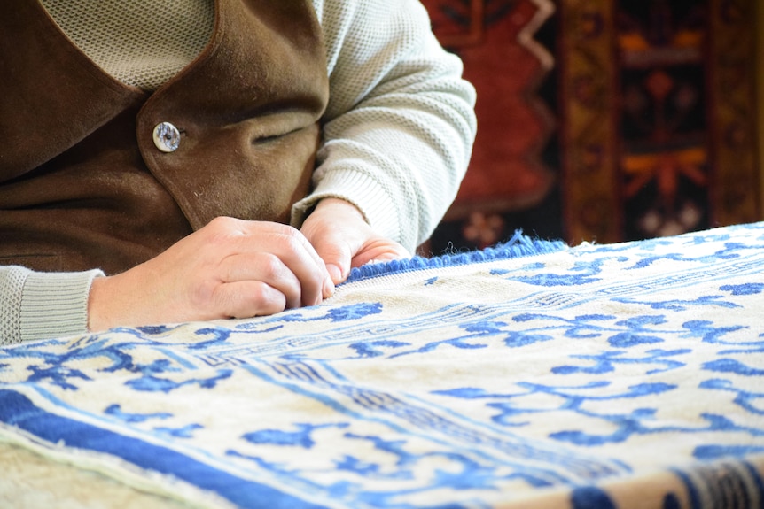 A man, face unseen, works on a carpet bearing an intricate design.