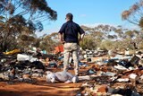 A man stands with his hands on his hips with his back to the camera surveying an area of scrub strewn with rubbish.