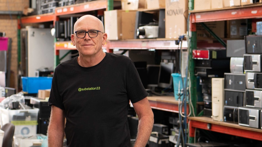 A bald man with black-rimmed spectacles smiles as he stands surrounded by computers and computer monitors.