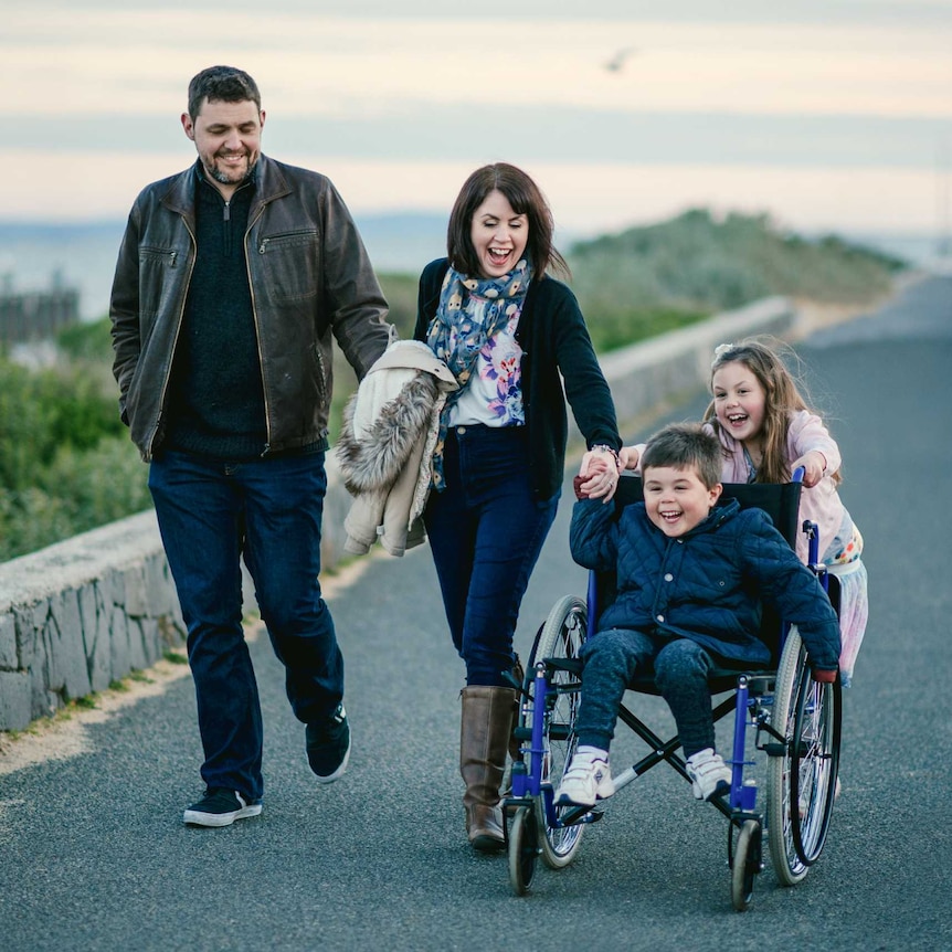 A man and a woman hold hands while walking alongside two children, one pushing the other in a wheelchair. They all smile.