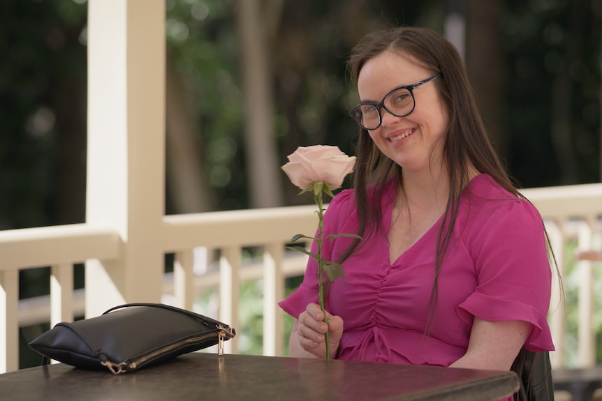 A woman wearing glasses and a pink shirt holds a flower.