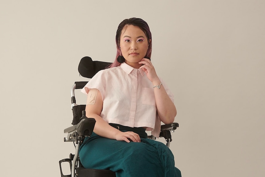 A young non-binary person of Asian background. They have pink highlights in their hair and are a wheelchair user