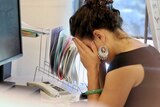 A woman puts her head in her hands while sitting at an office desk.
