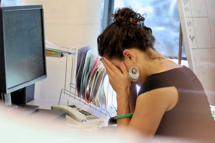 A woman puts her head in her hands while sitting at an office desk.