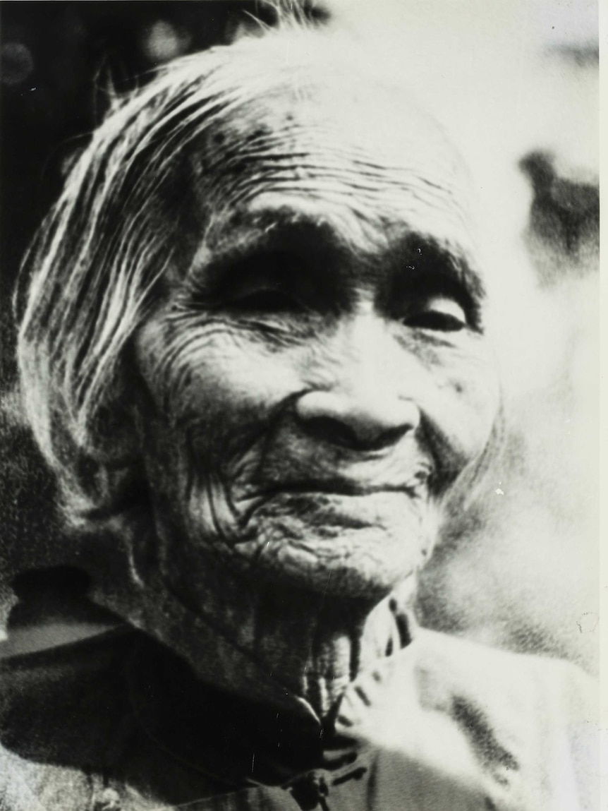 3:4 black & white portrait of elderly Chinese lady looking at the camera. Wry smile. Heavy wrinkles