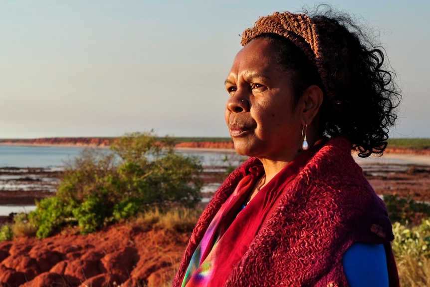 Portrait of a middle-aged Indigenous Australian woman looking out over a red landscape and water.