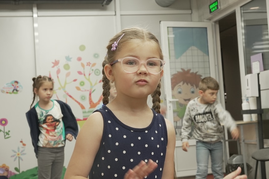 A young girl with braided pigtails and glasses with other children behind her. 