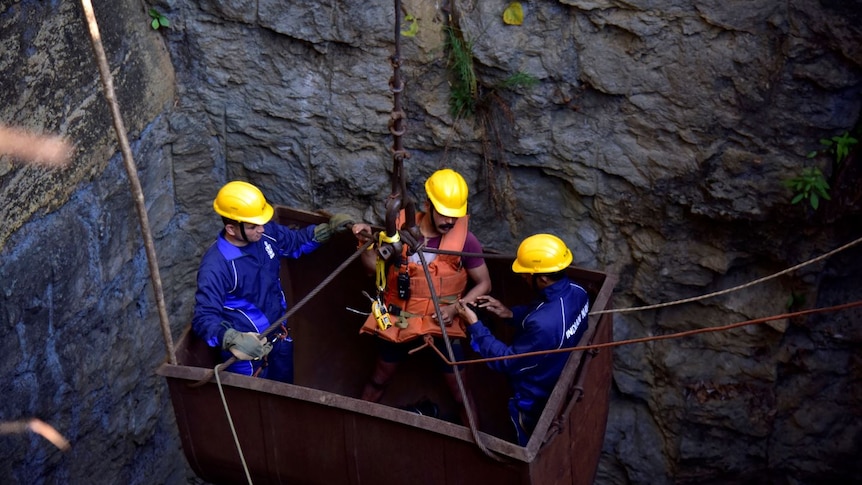 Three men in yellow hard hats are lowered into a large underground shaft in a large, rusted metal bucket.