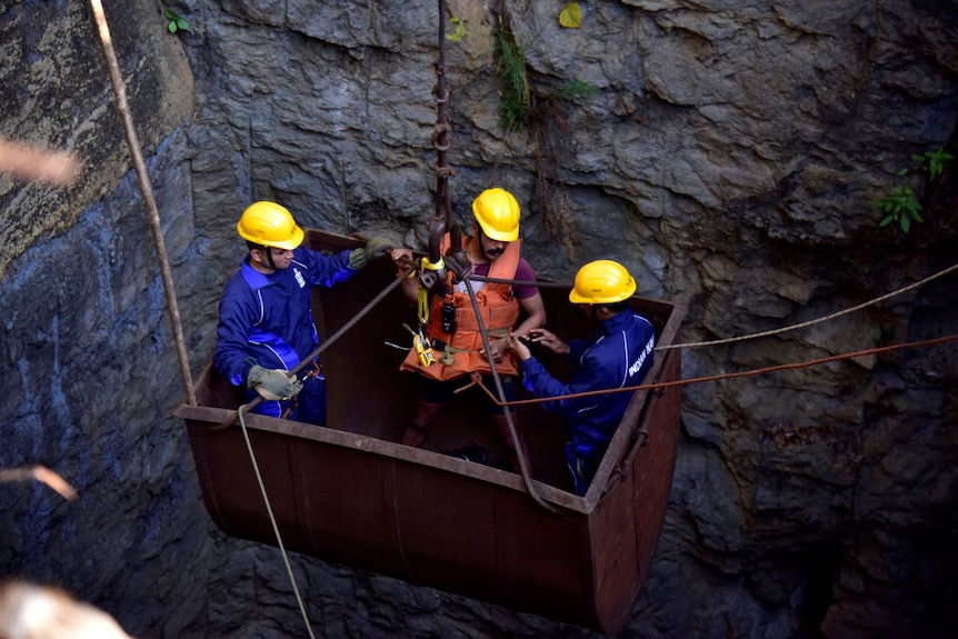Three men in yellow hard hats are lowered into a large underground shaft in a large, rusted metal bucket.
