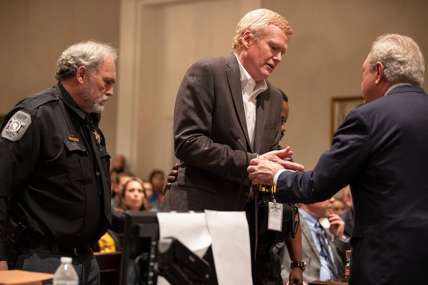 A middle white aged man in a suit is handcuffed in a courtroom with a uniformed guard behind him