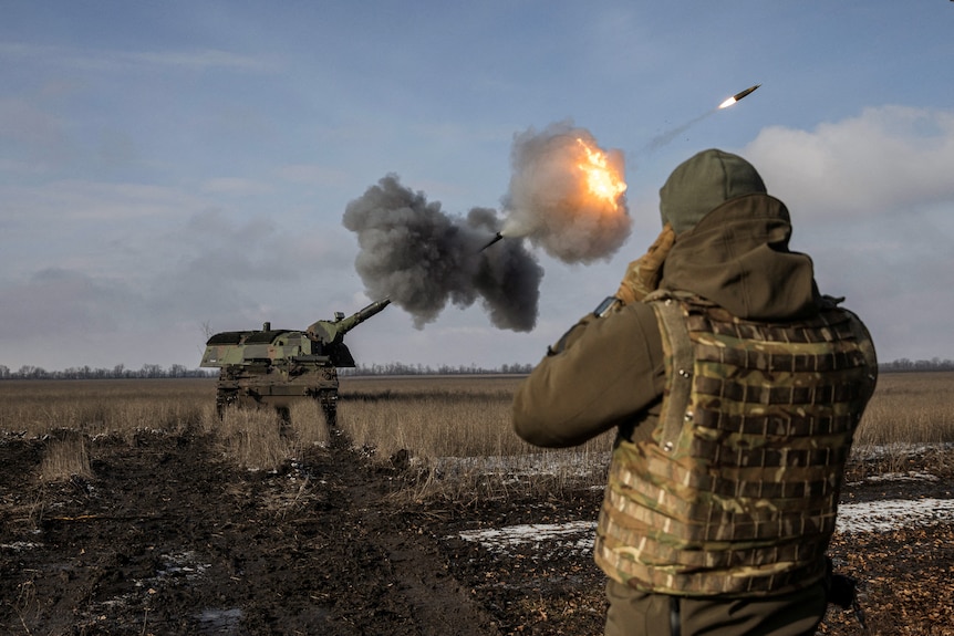 A soldier puts his hands over his ears as he watches a tank fire a missile into the sky.