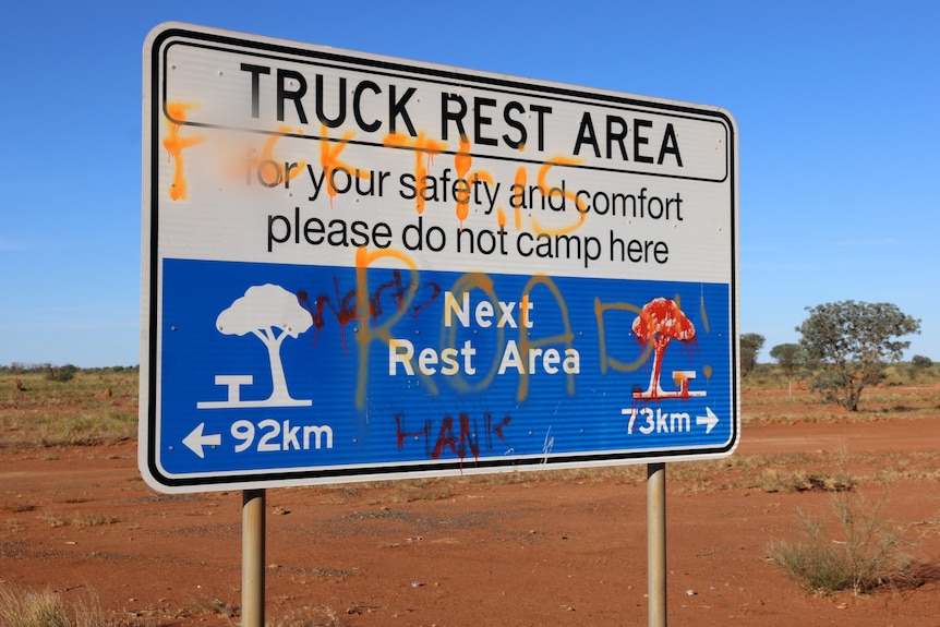 A graffitied rest stop sign along a dirt road.