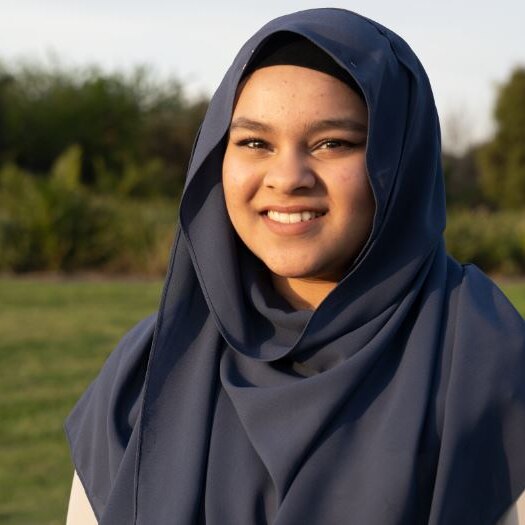 A teenage girl smiles and looks to the camera as she stands in a park during the day. She wears a blue head scarf.
