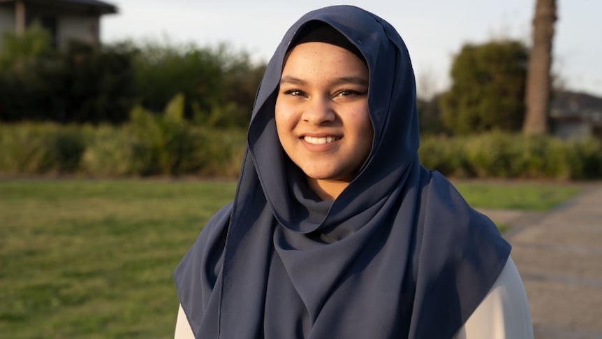 A teenage girl smiles and looks to the camera as she stands in a park during the day. She wears a blue head scarf.