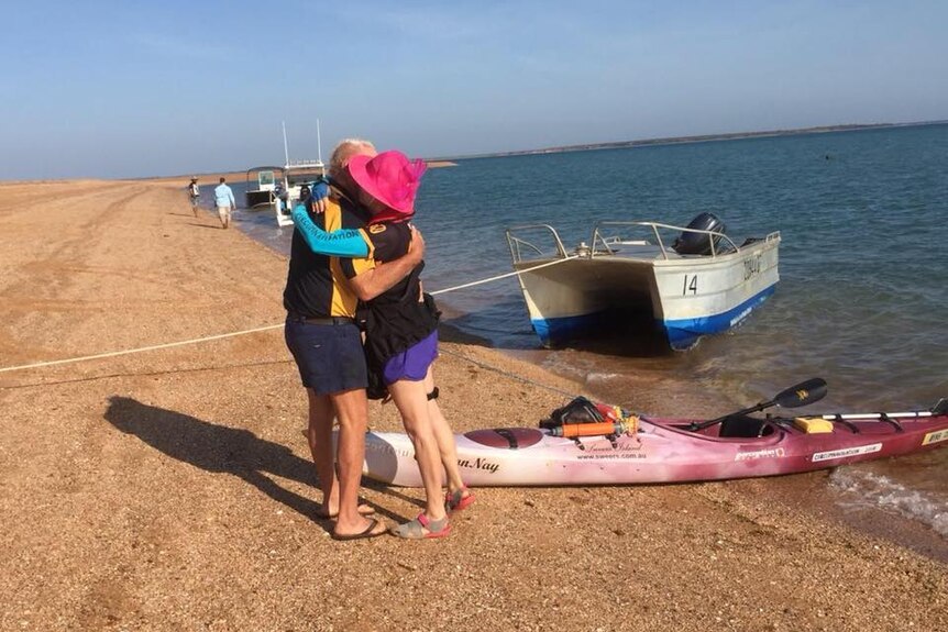 Un homme et une femme s'embrassent sur un littoral avec un bateau et un kayak derrière eux.
