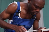 A man wearing a white singlet and blue overalls focuses intently as he bends over his work making a wooden frame
