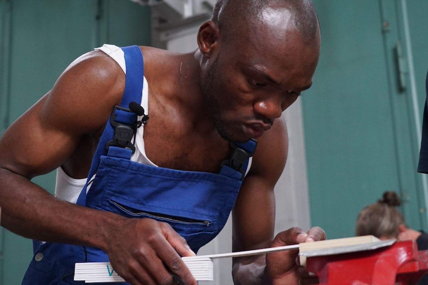 A man wearing a white singlet and blue overalls focuses intently as he bends over his work making a wooden frame