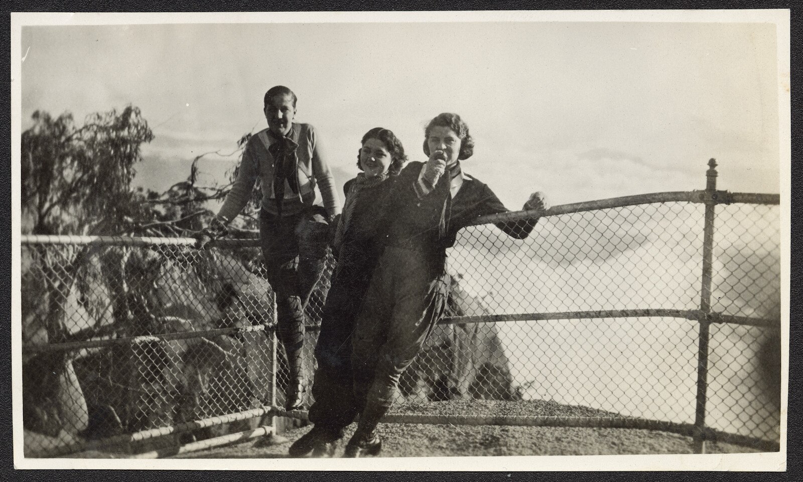 A black and white photo of a group of a young man and two young women leaning against railing on top of a rock lookout