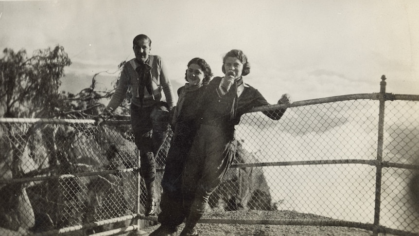 A black and white photo of a group of a young man and two young women leaning against railing on top of a rock lookout.