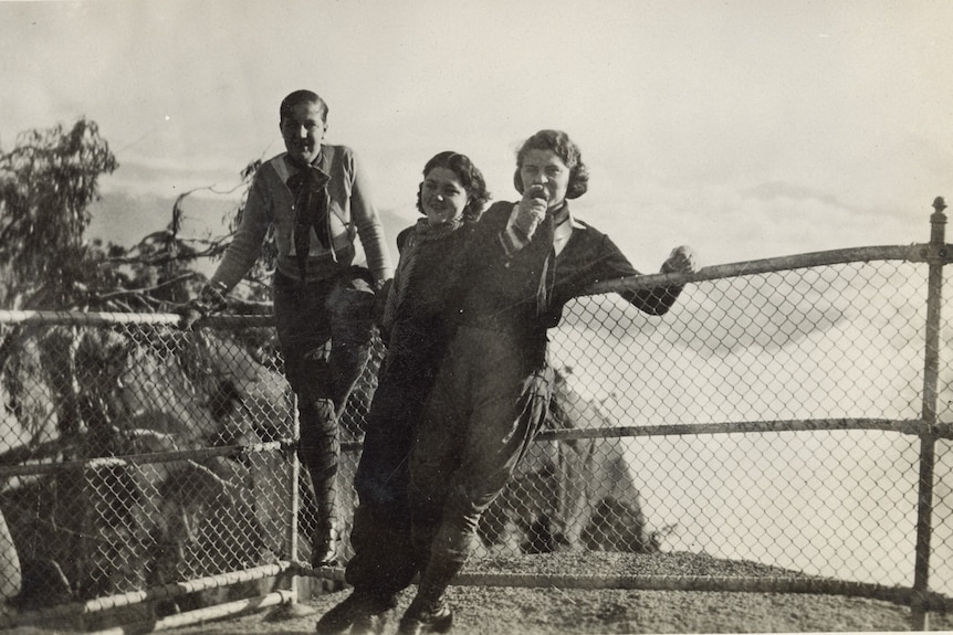 A black and white photo of a group of a young man and two young women leaning against railing on top of a rock lookout