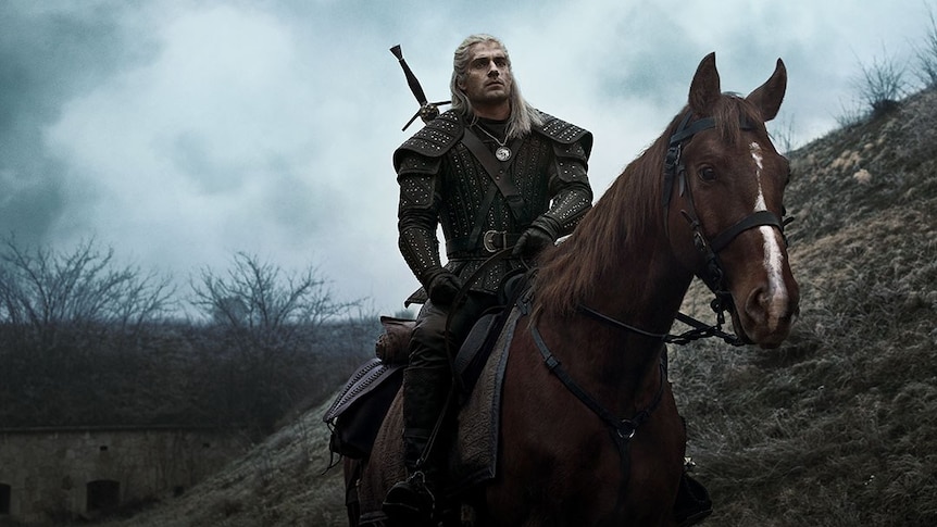 A man with long, blonde hair, rides a horse along a barren hill on a cloudy day