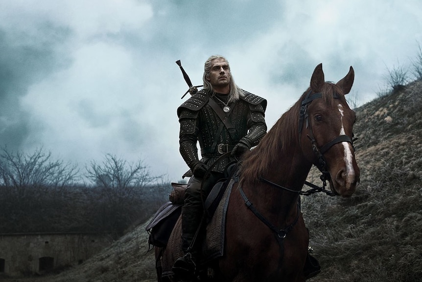 A man with long, blonde hair, rides a horse along a barren hill on a cloudy day