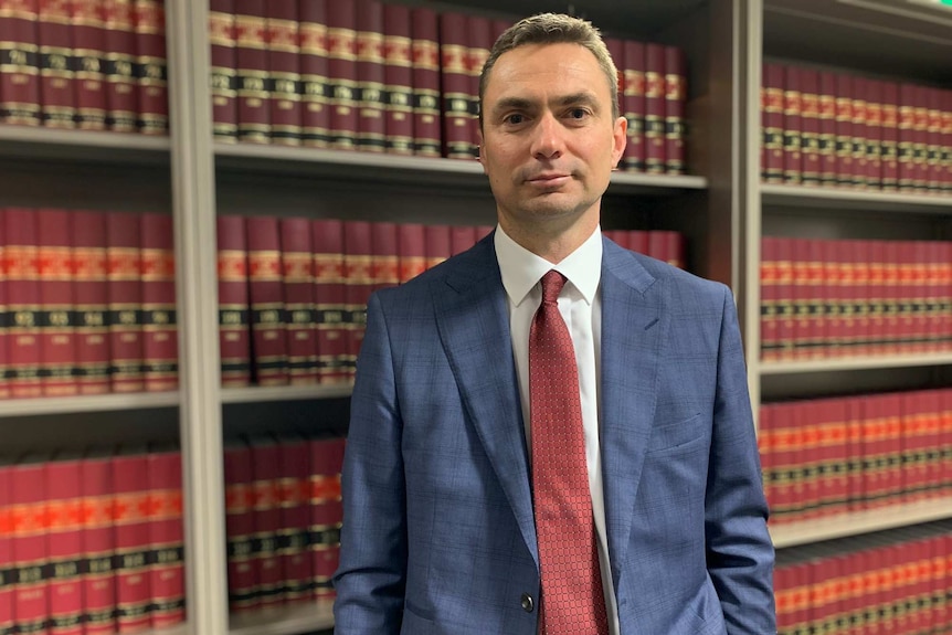 A man wearing a blue suit stands in front of a bookshelf of red and gold legal books.