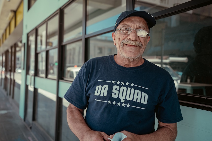 An elderly man in a t-shirt and cap poses against a row of offices. 