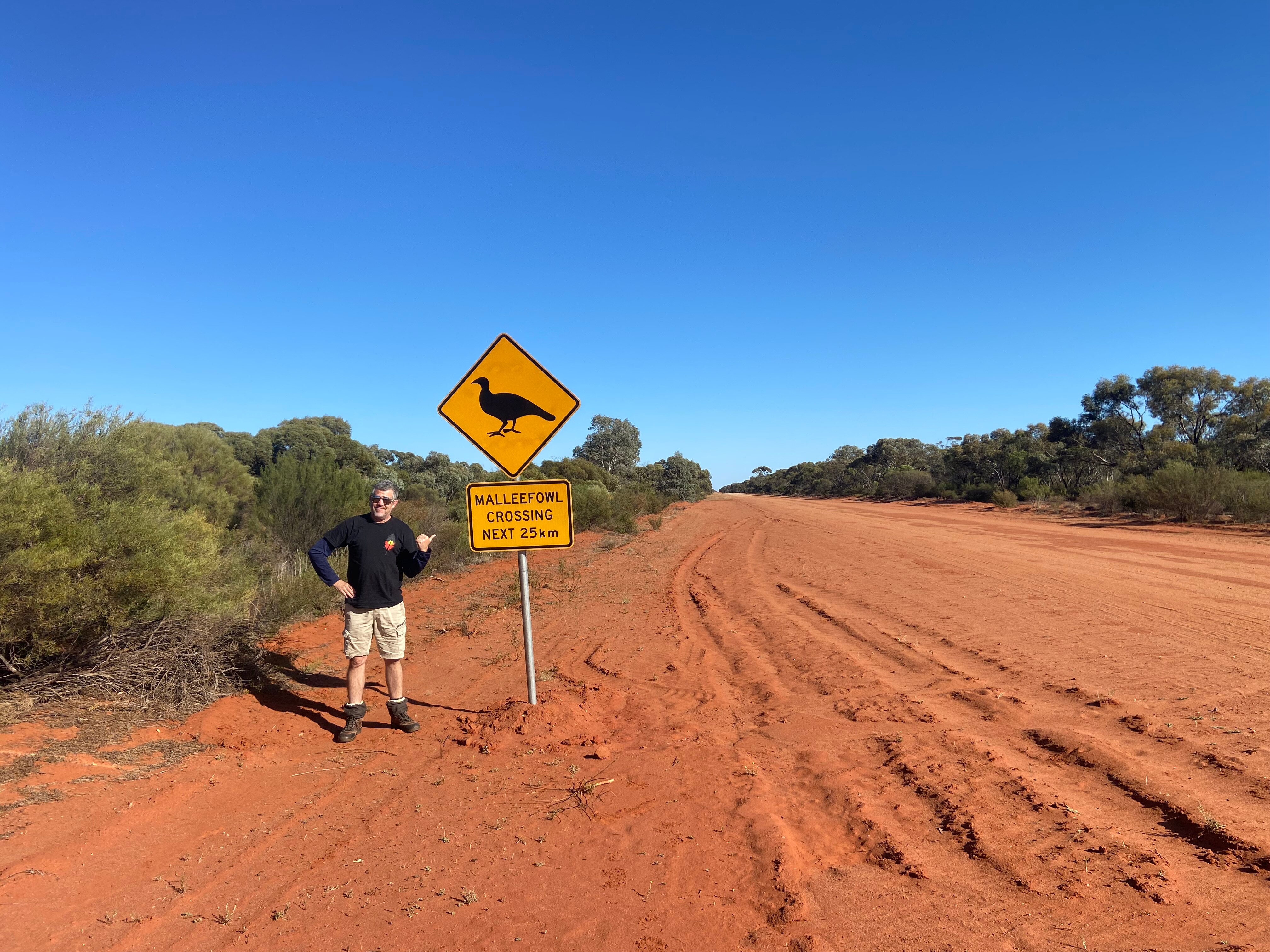 A man stands on a red dirt road next to a sign with a bird on it, and a blue sky.