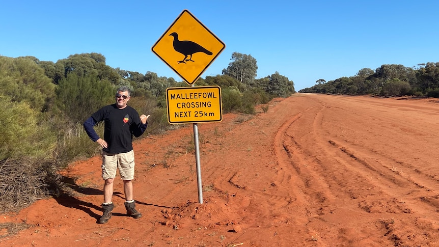 A man stands on a red dirt road next to a sign with a bird on it, and a blue sky.