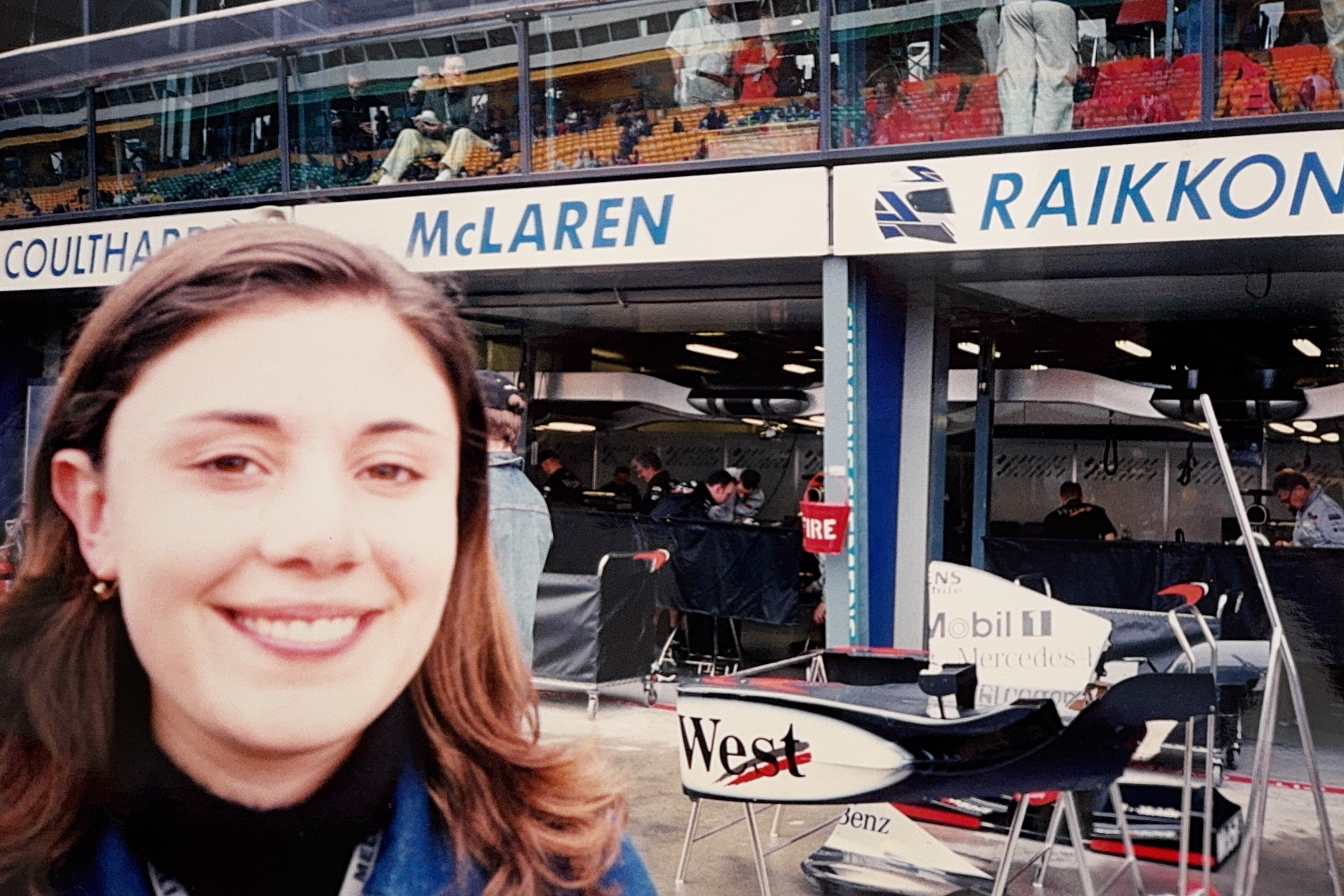 Woman with dark hair smiles standing in pit lane at car racing event. "McLaren" can be read on sign behind
