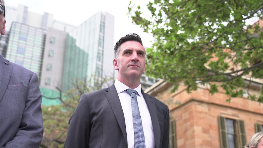 Fraser Ellis stares ahead with his hands behind his back, court buildings are in the background