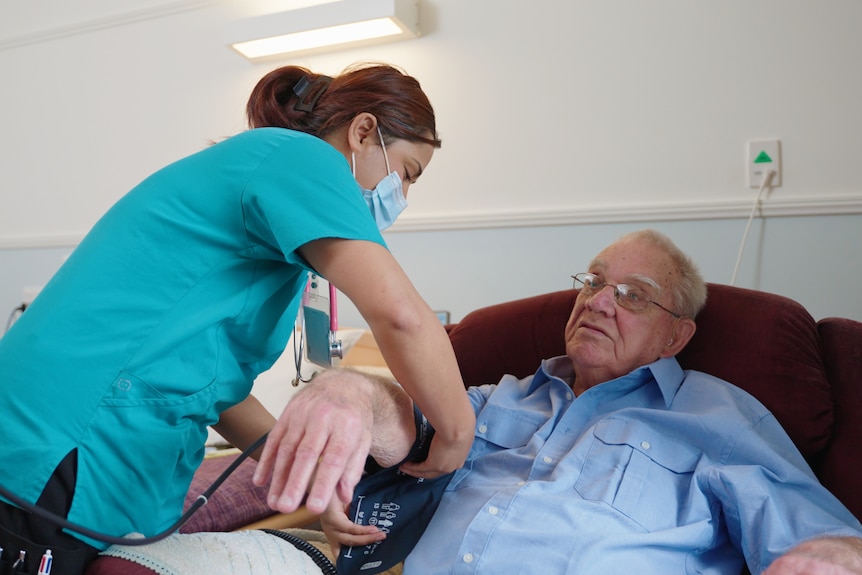 A female nurse attends to an elderly man in a bed.