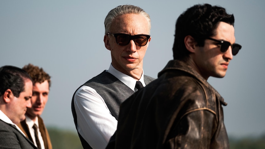 A film still of Adam Driver, with white hair and sunglasses, his arms folded, and Gabriel Leone, in a leather jacket and sunnies
