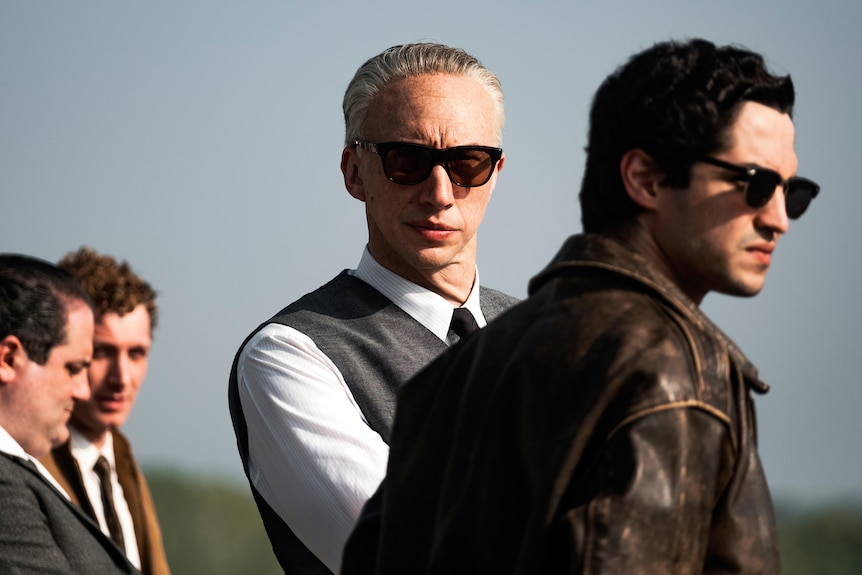 A film still of Adam Driver, with white hair and sunglasses, his arms folded, and Gabriel Leone, in a leather jacket and sunnies