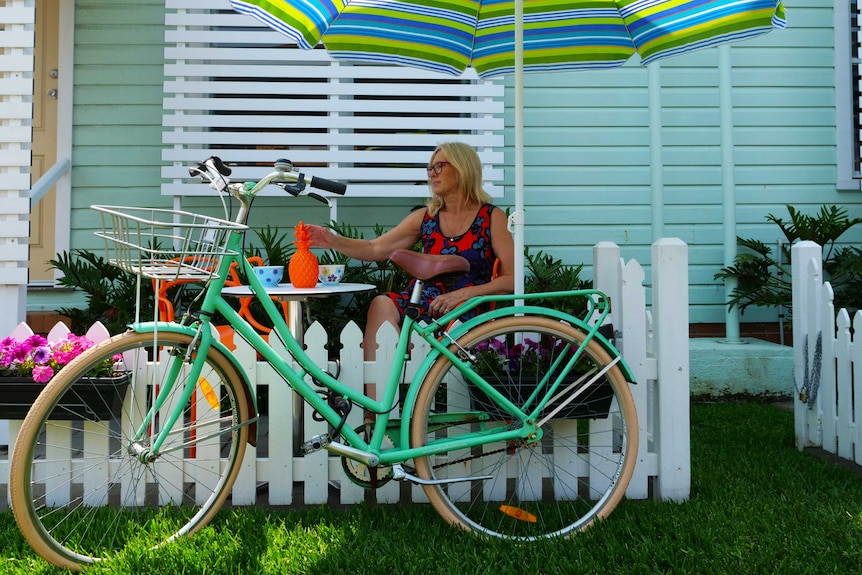 A woman sitting outside an older-style residence at a small round table, looking to the side. A retro bike is in front of her.