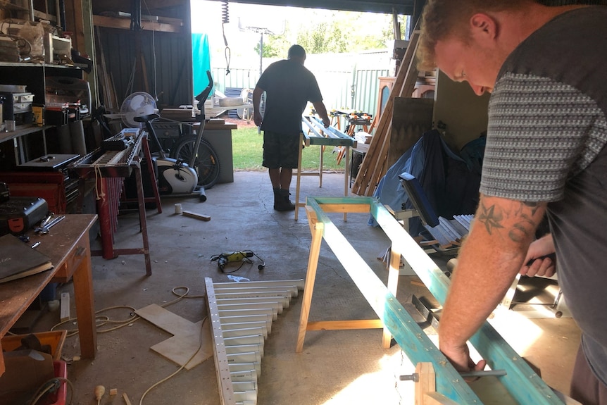 A man in a shed working on a blue wooden frame. Pipes sit on the floor. Another man in the background works on a second frame.