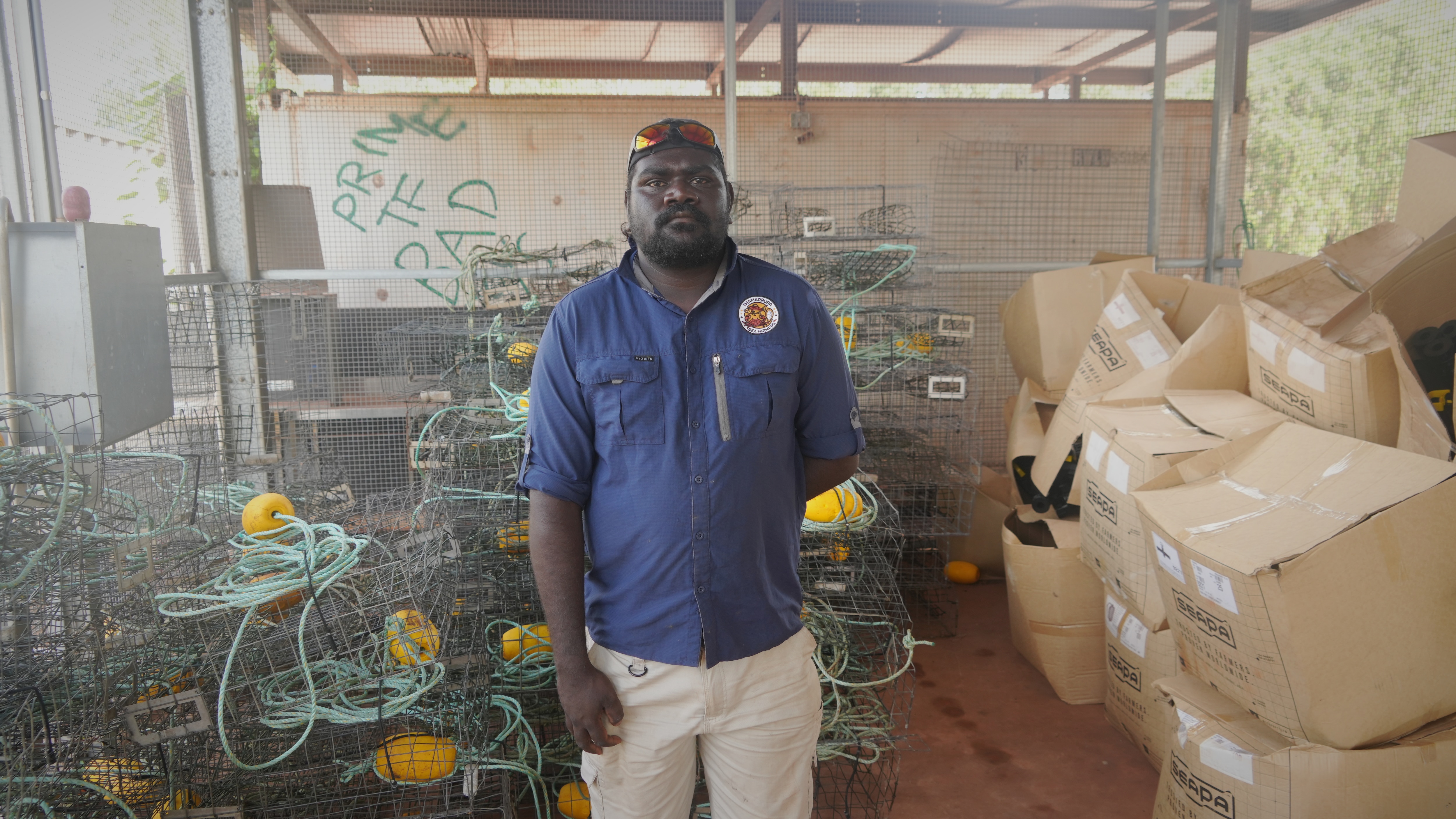 Indigenous man wearing a blue shirt and white pants standing in front of dozens of crab pots.
