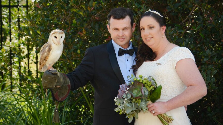 A husband and wife posing with a white barn owl and a bouquet of flowers