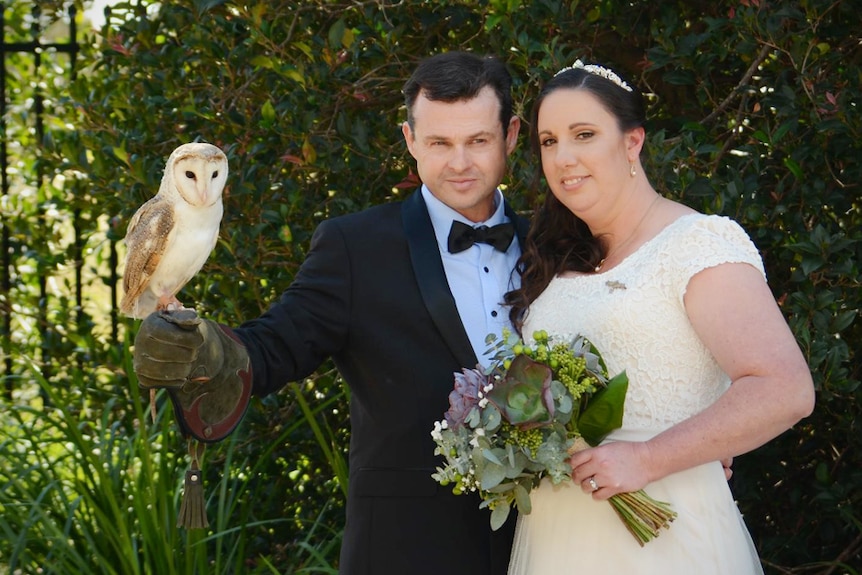A husband and wife posing with a white barn owl and a bouquet of flowers