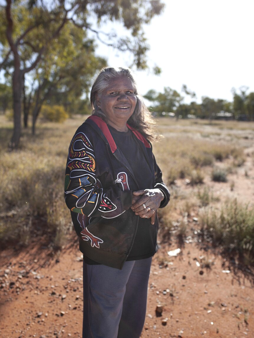 An Aboriginal woman wearing a black jacket smiles at the camera while standing in a bush setting.