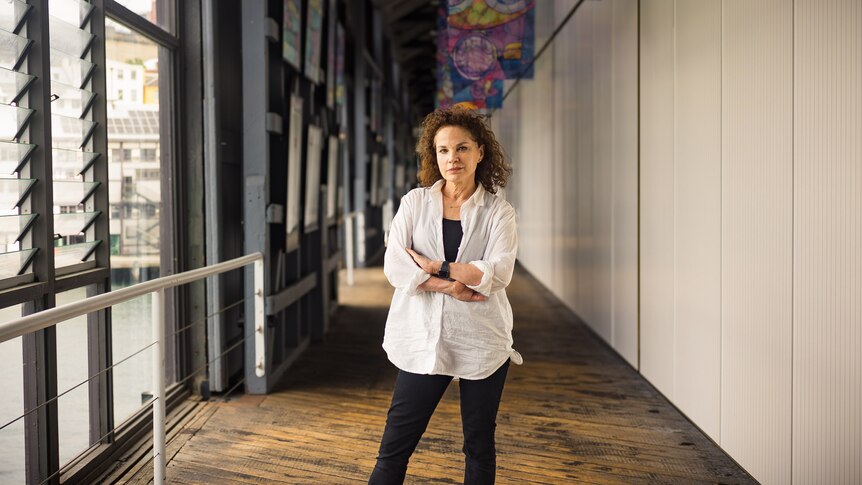 Sigrid Thornton, a 64-year-old white woman with curly brown hair wearing a white blouse and black pants standing in a corridor