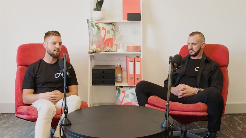 Two men sitting in red chairs speak into microphones.