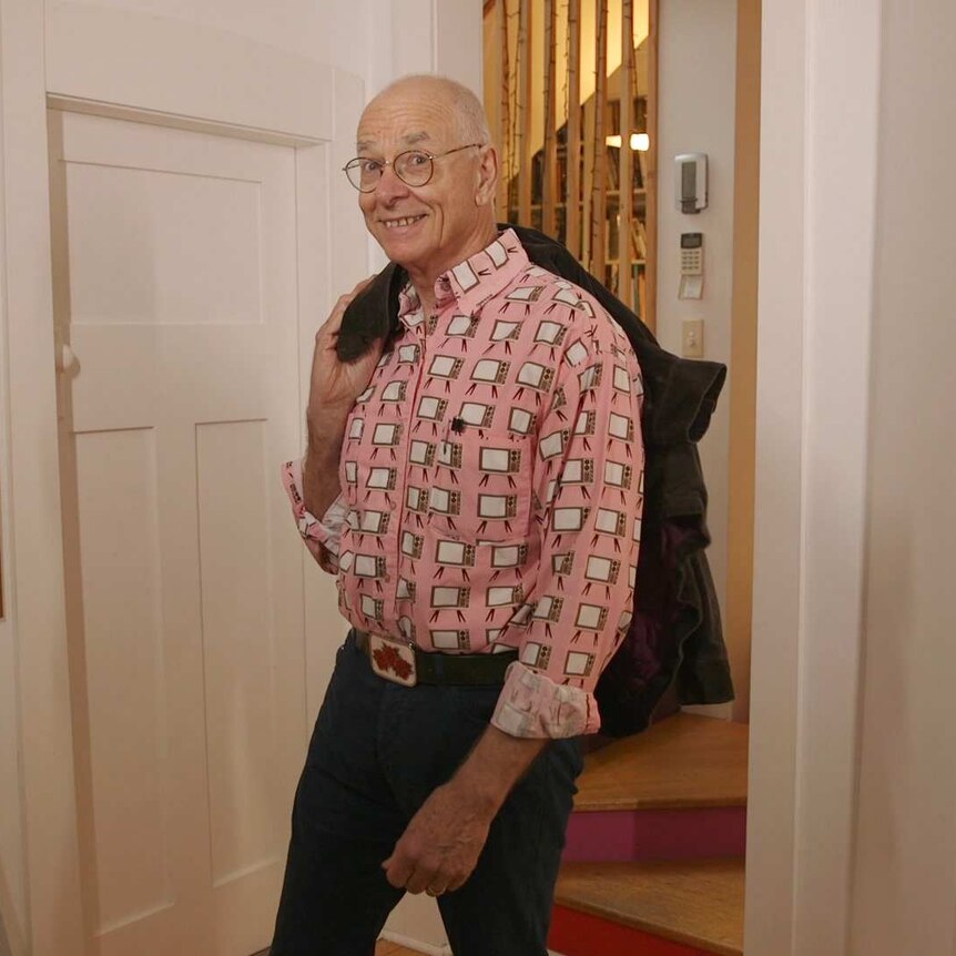Dr Karl smiles at the camera in a pink collared shirt with televisions across it, holding a coat over his shoulder.