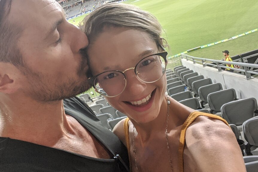 Erin Hill smiles while her husband kisses her at a football match.