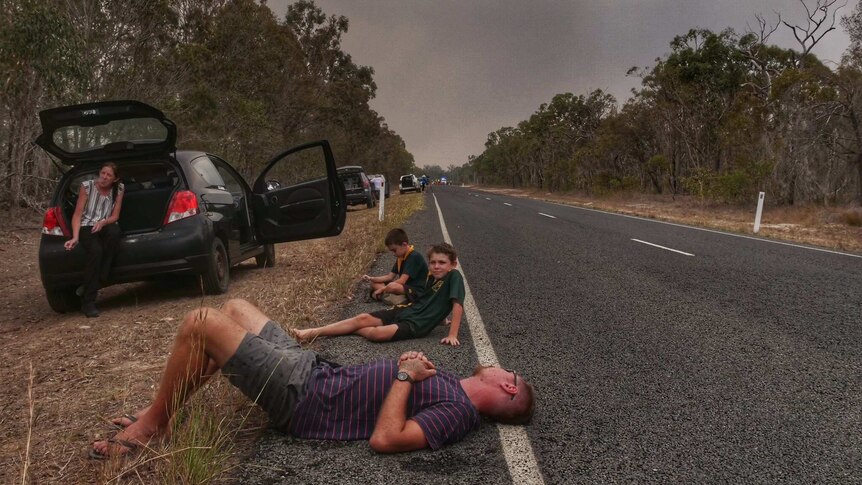 A man and children sit on the side of the road and cars are parked on the edge as smoke fills the air.