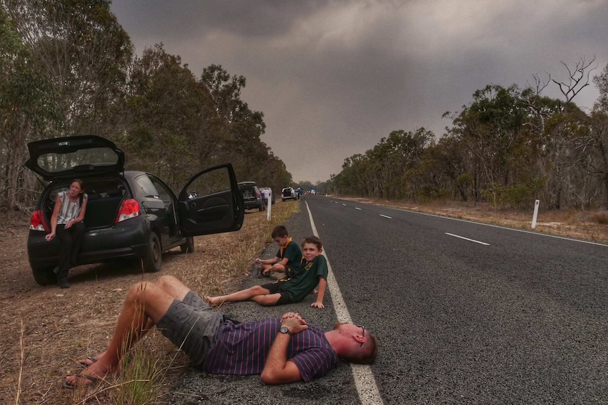 A man and children sit on the side of the road and cars are parked on the edge as smoke fills the air.