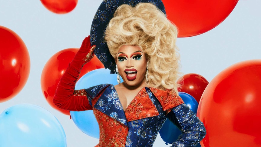 Drag queen Brita holds a large sparkly blue hat against her tall blonde wig. She wears a sparkling red, white and blue gown.